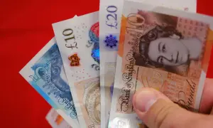 Dłoń trzyma banknoty: 5, 10 i 20 funtów. Różne nominały funtów widać na czerwonym tle.