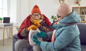 Dwoje starszych ludzi, ubranych w ciepłe czapki i szaliki, próbuje ogrzać się przy małym grzejniku. Wyglądają, jakby zamarzali w swoim domu w Wielkiej Brytanii.