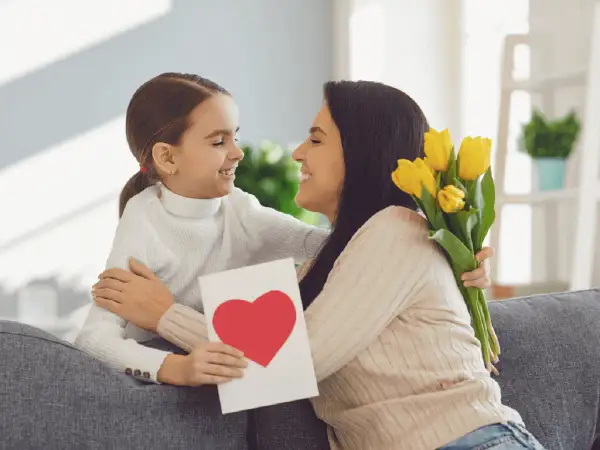 Uśmiechnięta dziewczynka wręcza mamie kartkę z sercem i bukiet żółtych tulipanów. Radosny Dzień Matki UK!