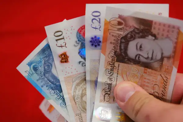 Dłoń trzyma banknoty: 5, 10 i 20 funtów. Różne nominały funtów widać na czerwonym tle.