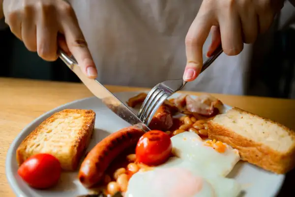Ręce krojące kiełbaskę na talerzu z fasolką, jajkiem sadzonym, pomidorkami i grzankami. To klasyczne angielskie śniadanie.